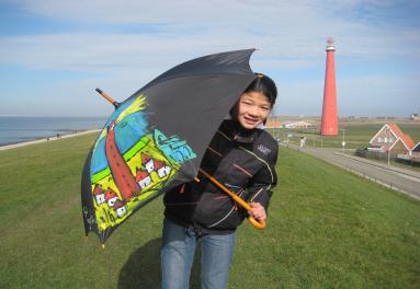 Foto van een persoon op een dijk met een paraplu in de hand waar een vuurtoren op staat afgebeeld. Op de achtergrond is de echte vuurtoren te zien.